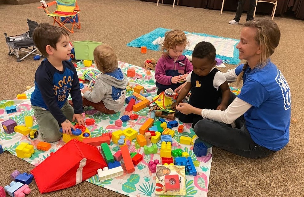 Image of an adult sitting on a carpet with four children and toys.