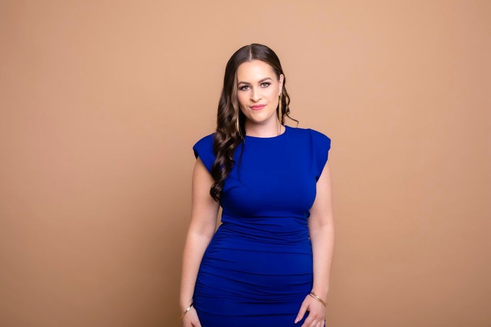 Photo of Courtney Stanley, standing in a blue dress against a beige background.