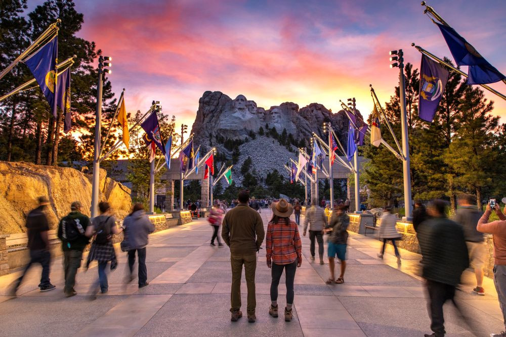 People standing in a walkway looking at Mt. Rushmore, with flags on the sides.