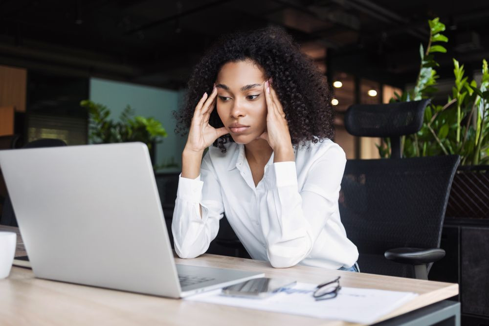 A business woman in a white blouse clasping her heads to her head in frustration, over a laptop.