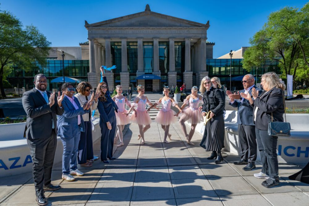 A crowd of people stand in front of a historic building, with ballet dancers, to celebrate cutting the ribbon.