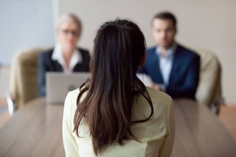 Image of two people interviewing a job candidate at a desk, with the candidate's back to the viewer.