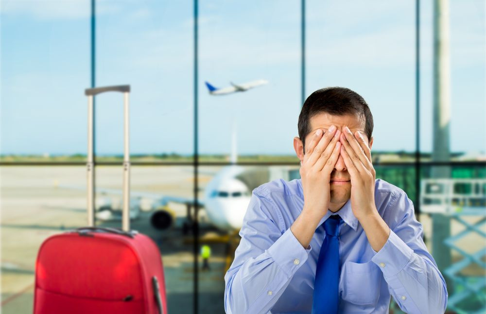 Photo of a man in an airport with his hands on his face, exasperated, with a red piece of carry on luggage next to him.